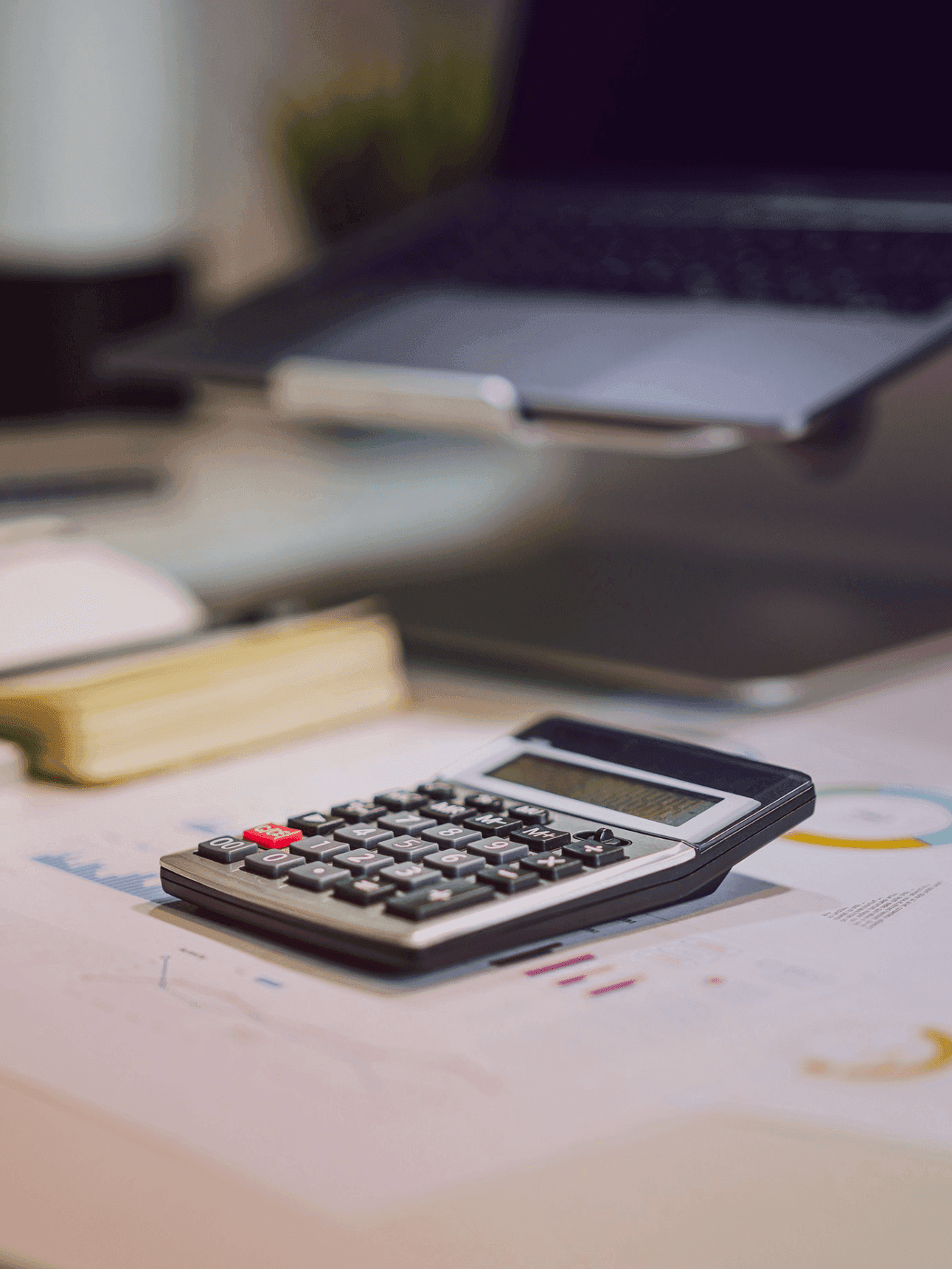 A calculator on a desk next to a laptop. Papers on the desk point to some kind of invoicing process