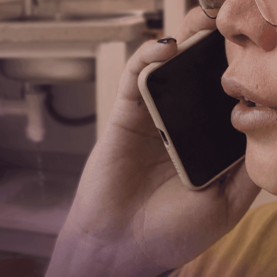 Person calling a plumber on a mobile while a pipe leaks under the sink behind them