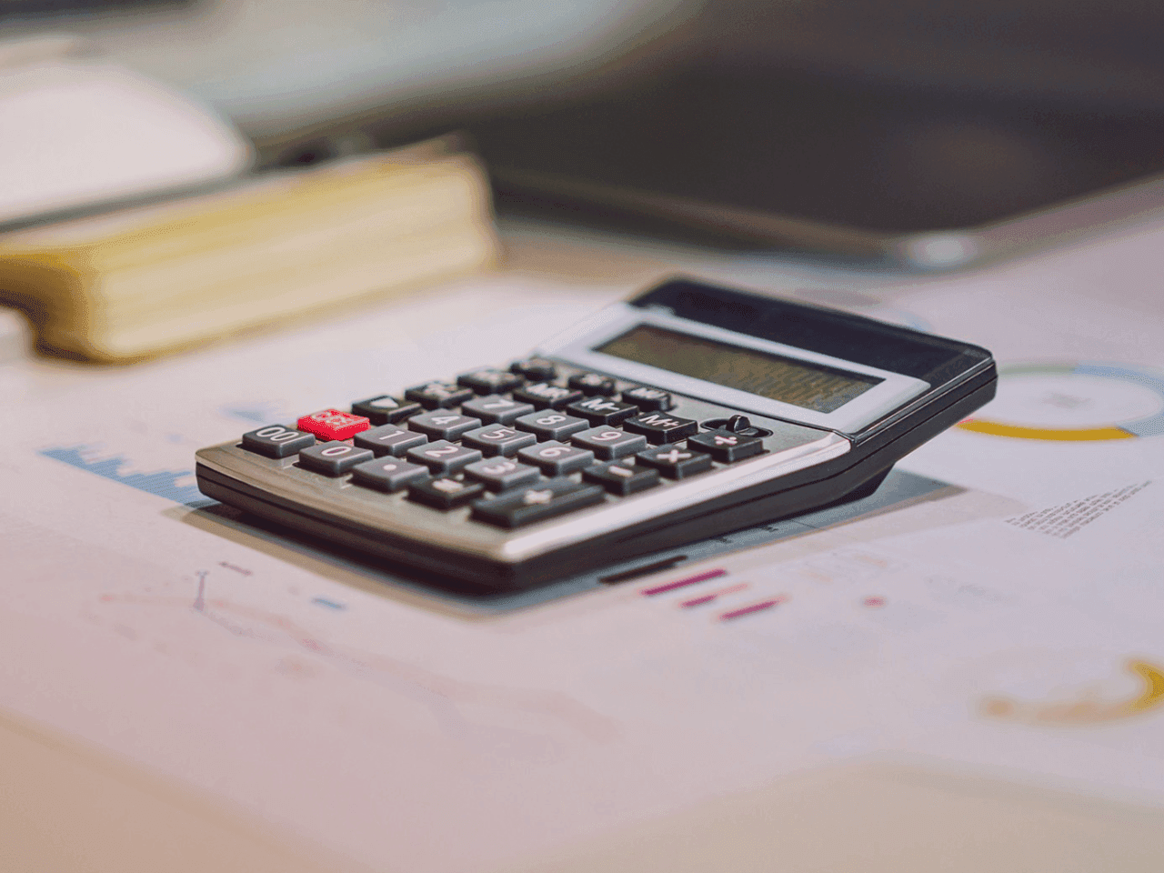 A calculator on a desk next to a laptop. Papers on the desk point to some kind of invoicing process