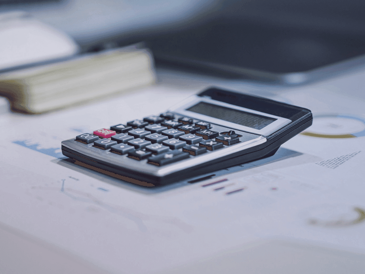 A calculator on a desk next to a laptop. Papers on the desk point to some kind of invoicing process