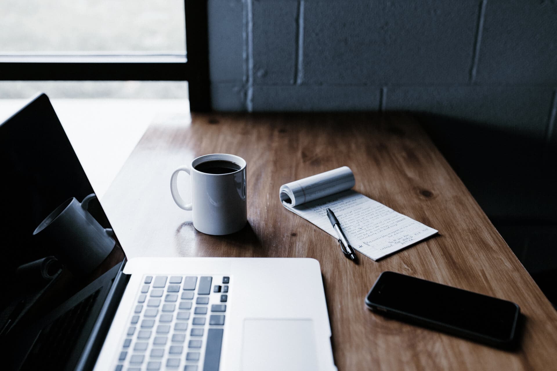 A laptop open on a wooden desk beside a coffee mug, a notepad with a pen, and a phone