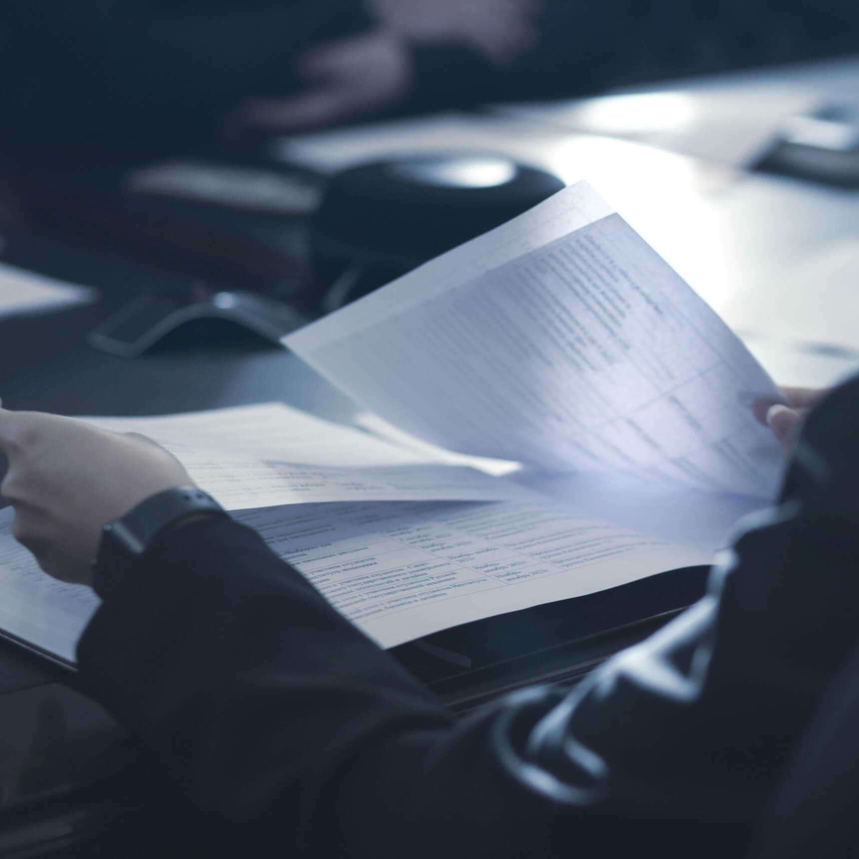 Hands turning the pages of a printed document on a meeting table, a speakerphone beside them