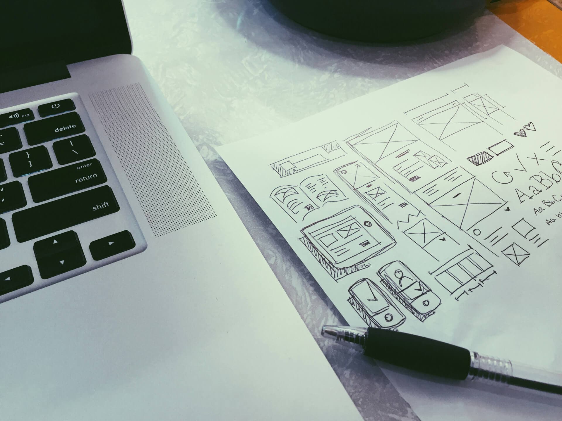 Hand-drawn website wireframe sketches on paper beside a laptop keyboard, with a pen resting on the page