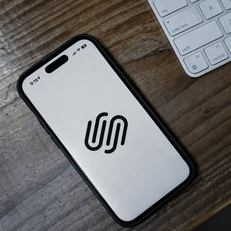 A smartphone showing the Squarespace logo, on a wooden desk beside a keyboard